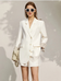 Woman wearing a white suit with sunglasses and holding a handbag against a light background
